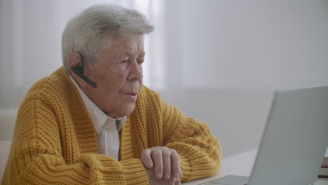 Ältere-Frau-Mit-Laptop-Computer-Ruft-Zu-Hause-Auf-Dem-Smartphone-An.-Alte-Frau-Führt-Einen-Videoanruf-Auf-Dem-Laptop,-Lächelt-Und-Redet-Glücklich-Drinnen-In-Einer-Gemütlichen-Wohnung.-Arzt-Videoanruf-älterer-Patient